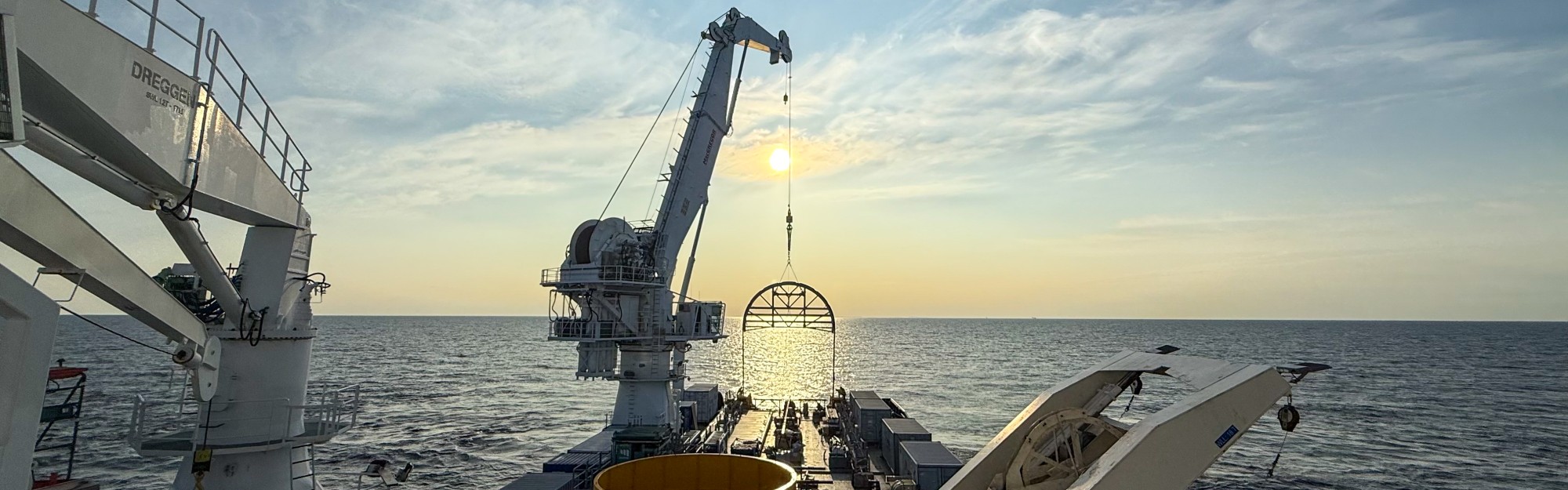 Crane lowering equipment into the ocean during sunset with a clear sky.