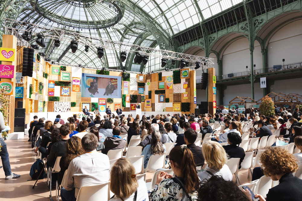 Conference at the ChangeNOW event, with four speakers discussing on stage in front of an audience, surrounded by colorful panels bearing inspirational messages.