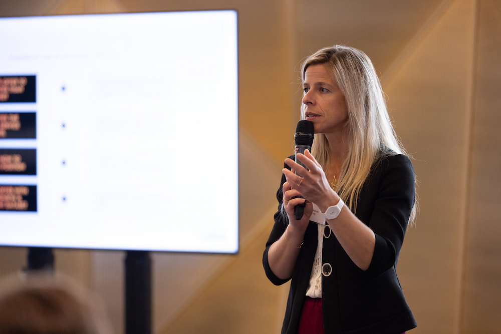 Speaker with long hair presenting at a conference, holding a microphone, engaging with the audience.